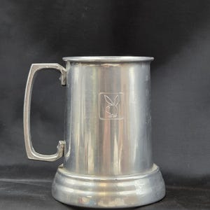 May include: A silver-colored metal beer mug with a large handle. The mug features a rectangular emblem with a rabbit logo. The base of the mug is a wider, circular shape. The mug is likely made of pewter or a similar metal.