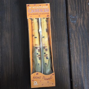 Puede incluir: Un par de velas esculpidas de 25 cm con forma de mazorcas de maíz con granos amarillos y hojas verdes, empaquetadas en una caja naranja con el texto 10" SCULPTURED CANDLES y Hand Painted! visible en el frente del empaque de plástico transparente.