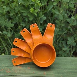 May include: A set of five orange plastic measuring cups stacked in a fan shape. Each handle features a circular hole at the end for hanging. The cups are labeled with volume measurements including 1/4 cup and 1/3 cup clearly visible on the handles.