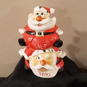 May include: A ceramic salt and pepper shaker set featuring two Santa Claus figures stacked on top of each other. The top Santa is red with a black belt and the word "SALT" on his belly. The bottom Santa winks and has the word "PEPPER".