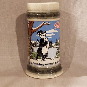 May include: A white ceramic mug with a black handle and the text "But First, Coffee" in a bold, black font. The mug is set against a blurred background, suggesting a cozy setting.