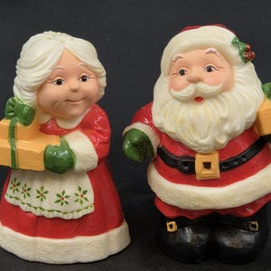 May include: A pair of Christmas figurines depicting Santa Claus and Mrs. Claus. Santa wears a red suit with a black belt and holds a gift. Mrs. Claus wears a red dress and holds a gift. Both figures have white hair and are smiling.
