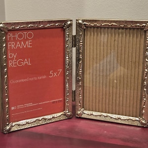 May include: A hinged, silver-colored photo frame by Regal, designed to hold two 5x7 inch photos. The frame has an ornate, decorative border and a red backing in one section. The other section has a cardboard backing.