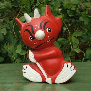 May include: A red ceramic devil figurine with white horns, eyes, and accents. The figurine has a grumpy expression and is sitting down. The devil's feet and hands are white. The figurine is on a green surface.