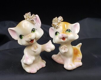 Puede incluir: Dos figuritas de gatitos de cerámica con detalles pintados. Un gatito es blanco con detalles rosas y el otro es marrón con detalles rosas. Ambos gatitos tienen ojos verdes y llevan una flor en la cabeza.