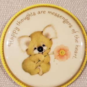 May include: Round, yellow-edged pin with a cream-colored center featuring a cartoon koala holding a flower. The koala is light brown with large ears and a black nose. The text around the edge reads, "Happy thoughts are messengers of the heart."