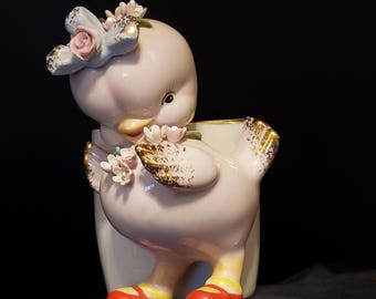 May include: A pink ceramic figurine of a duck wearing a blue and white flower crown and red shoes. The duck is holding a bouquet of pink flowers and is standing on a white pedestal.