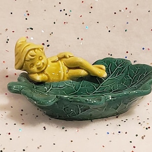 May include: A ceramic figurine of a yellow gnome resting in a green leaf-shaped dish. The gnome is lying down with its arms crossed. The dish has detailed leaf veins. The background is white with colorful confetti.