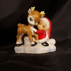 May include: A ceramic figurine featuring a deer and a child in a red and white outfit, hugging. The deer is brown with yellow antlers and white spots. The child has green eyes and is wearing a red coat and white hat. The base is white with a blue tint.