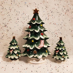 May include: Three ceramic Christmas trees in varying sizes. The trees are dark green with white accents, red and yellow ornaments, and gold star toppers. The largest tree has a candy cane and stocking decoration.