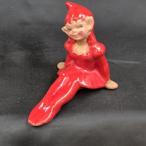 May include: A ceramic figurine of a seated elf with a red outfit and a pointed hat. The elf has a smiling face with closed eyes and tan skin. The figurine is on a black surface.