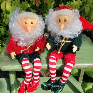 May include: Two plush Christmas elves with white beards, red and green outfits, and striped stockings are sitting on a green bench.