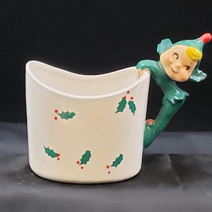 May include: White ceramic mug with a green elf figurine handle. The mug has a green holly leaf design with red berries.