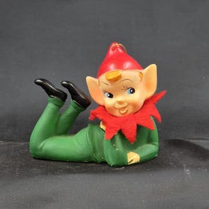 May include: A ceramic Christmas elf figurine in a reclining pose. The elf wears a green outfit, a red hat, and a red collar. The elf has black shoes, blue eyes, and a smiling expression. A gold string is attached to the hat.
