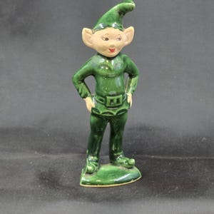 May include: A ceramic figurine of a green elf with a pointed hat, large ears, and a smiling face. The elf is wearing a green outfit with a belt and is standing with hands on hips. The figurine is on a green base.