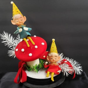 May include: Two vintage Christmas elves, one sitting on a red and white mushroom and the other sitting on a black base with white snow. Both elves are wearing yellow hats with gold glitter and have closed eyes.