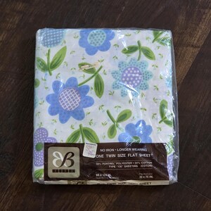 May include: A new, packaged twin-size flat sheet with a floral pattern in blue, purple, and green on a white background. The packaging includes text that reads "NO IRON - LONGER WEARING" and "ONE TWIN SIZE FLAT SHEET". The sheet measures 66 x 104 inches.