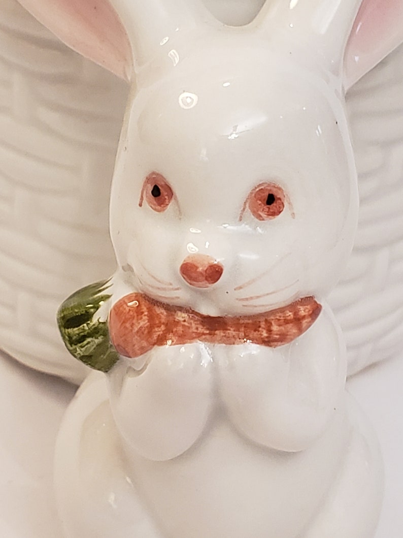以下が含まれることがあります： 白い陶器製のウサギの置物で、足にニンジンを持っています。ウサギはピンクの耳と茶色の目を持っています。