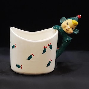 May include: White ceramic mug with a green elf handle and holly leaf design. The elf is peeking over the rim of the mug.