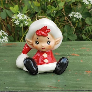 May include: A vintage ceramic Christmas elf ornament. The elf is wearing a red and white outfit with black shoes. The elf has a white hat with a red accent and a sweet expression. The ornament is sitting on a green surface.