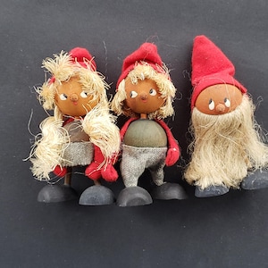 May include: Three wooden dolls with red hats and white hair. The dolls have painted faces with black eyes and brown skin. The dolls are standing on a black surface.