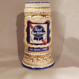 May include: A ceramic Pabst Blue Ribbon beer stein. The stein is beige with blue accents and features the Pabst Blue Ribbon logo in a blue and red design. The words "Pabst Brewing Company" are also visible.