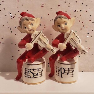May include: A pair of vintage Christmas elf figurines. Each elf is dressed in a red outfit and hat, playing a white violin. They sit atop white salt and pepper shakers with musical notes and the letters "S" and "P".