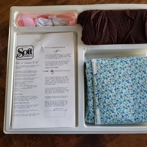 May include: A craft kit for making a soft sculpture. The kit includes a printed instruction sheet, a spool of brown yarn, two spools of white yarn, a piece of pink ribbon, and a piece of blue fabric with a floral pattern.