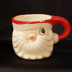 May include: A ceramic mug shaped like Santa Claus. The mug features a white face and beard, a red hat and handle, and a pink nose. The mug is designed with a festive theme, perfect for holiday beverages.
