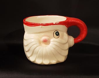 May include: A ceramic mug shaped like Santa Claus. The mug features a white face and beard, a red hat and handle, and a pink nose. The mug is designed with a festive theme, perfect for holiday beverages.
