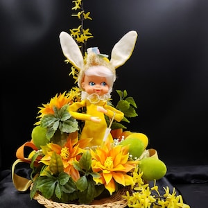 May include: A yellow bunny doll with white ears sits in a wicker basket filled with yellow and green flowers. The doll is wearing a yellow dress and has a white face with painted blue eyes.