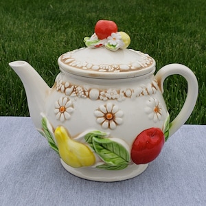 May include: White ceramic teapot with a floral design and fruit accents. The teapot has a lid with a red cherry and yellow flower on top. The teapot is decorated with pears, apples, and leaves.