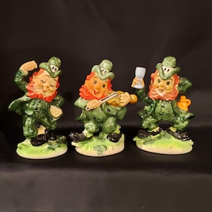 May include: Three ceramic leprechaun figurines. Each figure is wearing a green suit and hat, with a red beard and smiling face. One is dancing, one playing a violin, and one holding a glass.
