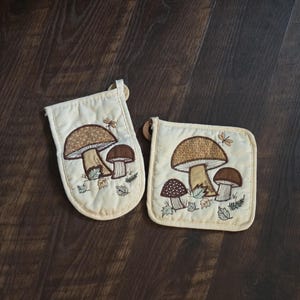 May include: A cream-colored quilted oven mitt and a square pot holder, both featuring embroidered brown mushroom designs with small green leaves and foliage accents. Each piece includes a small fabric loop for hanging on the top corner.
