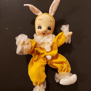 May include: A vintage doll dressed in a yellow jumpsuit with white ruffled trim at the wrists and ankles. The doll has a bunny-shaped hood, a painted face with rosy cheeks, and long, upright ears. The doll's shoes are white.