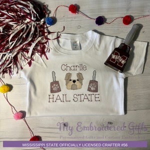 May include: White t-shirt with embroidered brown dog, two bells, and the text "Charlie" and "Hail State". A red bell with a white "M State" logo is on the right side of the shirt.