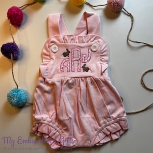 May include: Pink baby romper with a ruffled neckline and leg openings. The romper has a white embroidered monogram with the letters "AJ" and two brown bunnies.