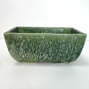 以下が含まれることがあります： 側面が織り模様のようなテクスチャ加工が施された、長方形の緑色のセラミックプランター。プランターは斑点模様の仕上がりで、わずかに湾曲した形状をしています。全体的なデザインは、屋内または屋外での使用に適した、自然で有機的な美しさを連想させます。