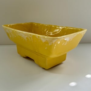 Pode incluir: Um vaso de cerâmica amarelo com formato retangular e uma borda ligeiramente ondulada. O vaso tem quatro pequenas pernas.