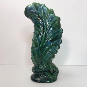 May include: A tall, decorative ceramic vase in the shape of a stylized leaf. The vase is glazed in shades of green and blue, with a glossy finish. The base is a dark green, and the overall design is reminiscent of Art Deco style.