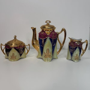 May include: A vintage tea set comprising a teapot, sugar bowl, and creamer. The set has gold trim and accents, with a cream, blue, and burgundy colour scheme. The teapot is approximately 20 cm tall.