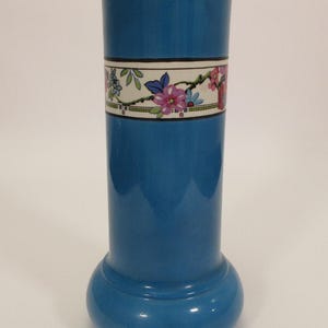 May include: A cylindrical blue ceramic vase with a flared rim and a rounded base. A horizontal white band encircles the upper section, featuring a colorful floral pattern with pink, blue, and green botanical details outlined in black.