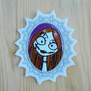 Könnte beinhalten: Gestickter Aufnäher mit einem Cartoon-Mädchen mit braunen Haaren und einem lila Hintergrund. Das Mädchen hat große Augen, einen gestickten Mund und einen weißen Spitzenrand um sie herum. Der Aufnäher ist weiß mit einem silbernen Spitzendesign.