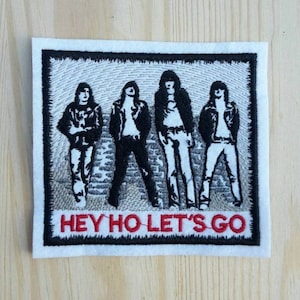 Può includere: Toppa ricamata in bianco e nero con quattro figure di una band, con il testo "HEY HO LET'S GO" in rosso.