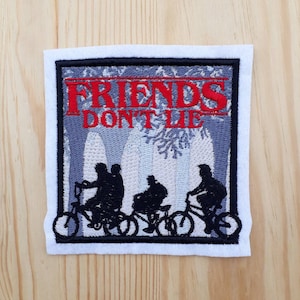 Könnte beinhalten: Gestickter Aufnäher mit grauem Hintergrund, auf dem eine Gruppe von vier Personen auf Fahrrädern zu sehen ist. Der Text "FRIENDS DON'T LIE" ist in roten Buchstaben über den Figuren.