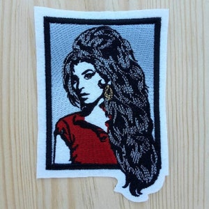 May include: Embroidered patch featuring a portrait of a woman with long black hair, wearing a red shirt and a gold earring. The patch is framed with a black border and set against a white background.