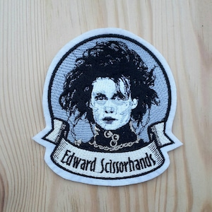 Può includere: Toppa ricamata con un'immagine in bianco e nero di Edward mani di forbice con il testo "Edward Scissorhands" su uno stendardo bianco.