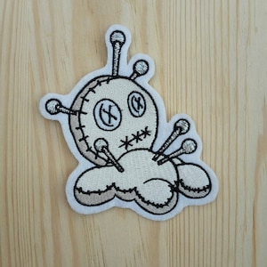 May include: White embroidered patch of a voodoo doll with black stitching and silver pins. The doll has a stitched-on mouth and eyes.