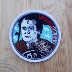 Può includere: Toppa ricamata con Bill Murray nei panni di Phil Connors dal film Ricomincio da capo. La toppa è circolare e ha uno sfondo bianco con un cielo blu e nuvole. Il testo "Groundhog Day" è scritto in lettere rosse e blu intorno al bordo della toppa.
