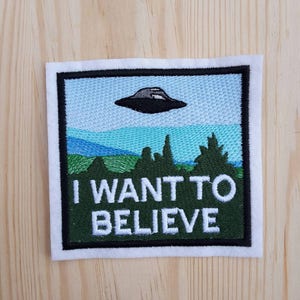 Può includere: Toppa ricamata con sfondo verde e testo bianco che recita "I WANT TO BELIEVE". Un UFO nero vola sopra un paesaggio verde con alberi.